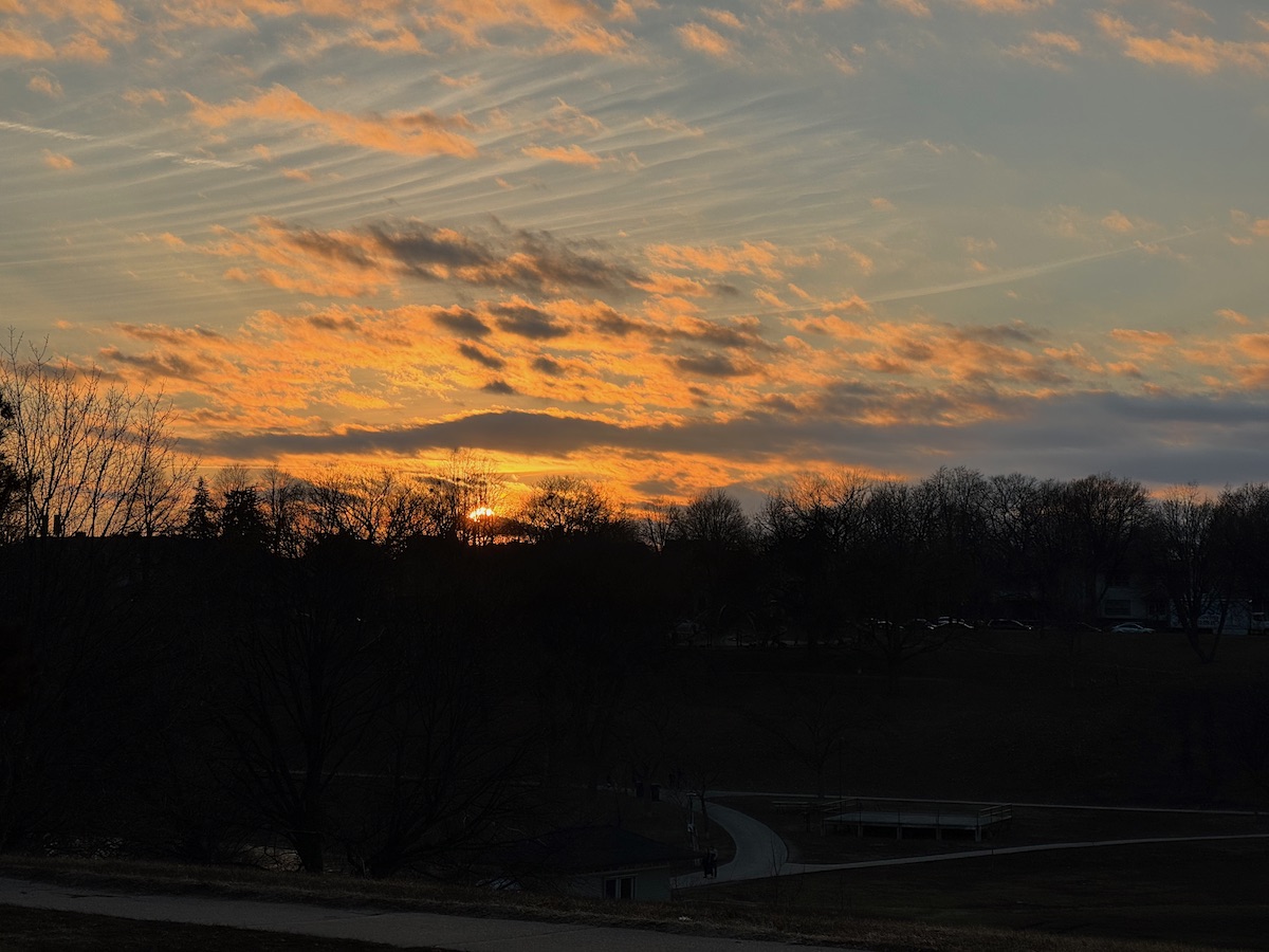 A photo of the sunset over Powderhorn Park in Minneapolis, MN. The treeline is about halfway vertically, with everything below it dark. Above the treeline, the sky surrounding the setting sun is dappled with clouds, varying from yellow, orange and a bit of pink. Farther from the sun, the sky is light blue with less clouds.