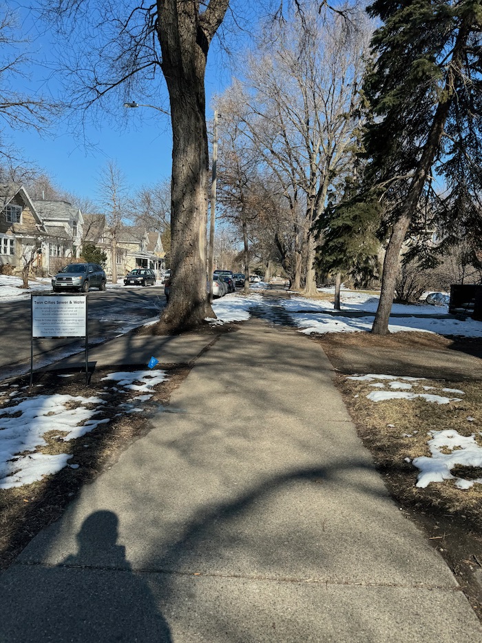 A photo of a sidewalk in the Como Neighborhood in Northeast Minneapolis, MN. There are traces of snow in the foreground yards and more snow farther in the background. The trees are leafless and there are some cars parked in the street, with a few houses visible on the left side.