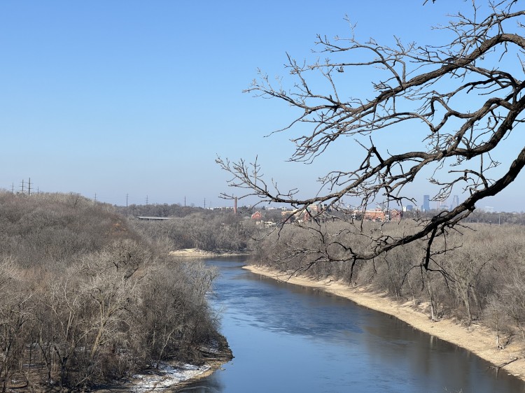 A photo of the Mississippi River from the walking path along Mississippi River Boulevard in St. Paul, MN. Both sides of the river have leafless trees and sand along the river. In the distance, the skyline of Minneapolis is visible through tree branches in the foreground breaking in from the right side of the picture.